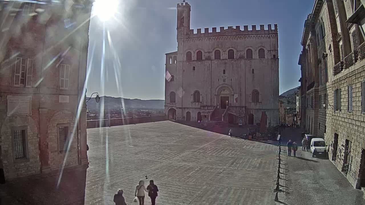A large, light-colored stone building stands in a sun-drenched, paved plaza, flanked by other buildings, under a bright, clear sky.