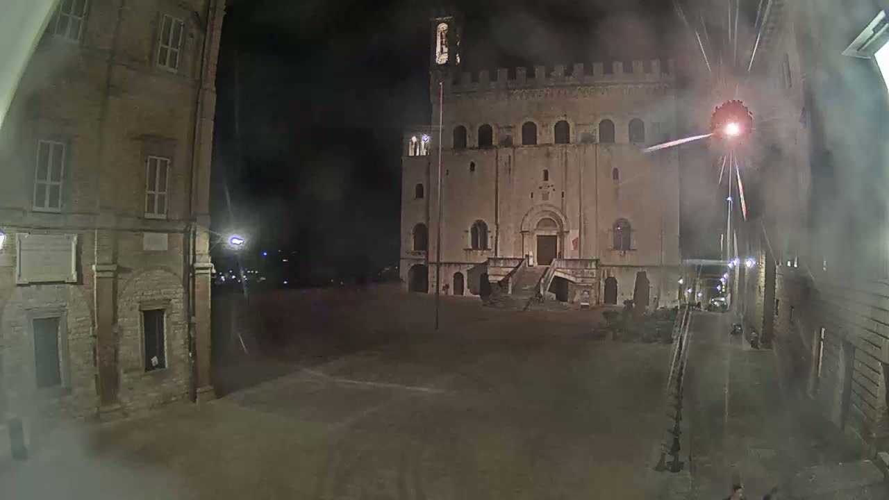 A large stone building with a staircase stands in a dimly lit, empty square at night, with fireworks visible in the sky.