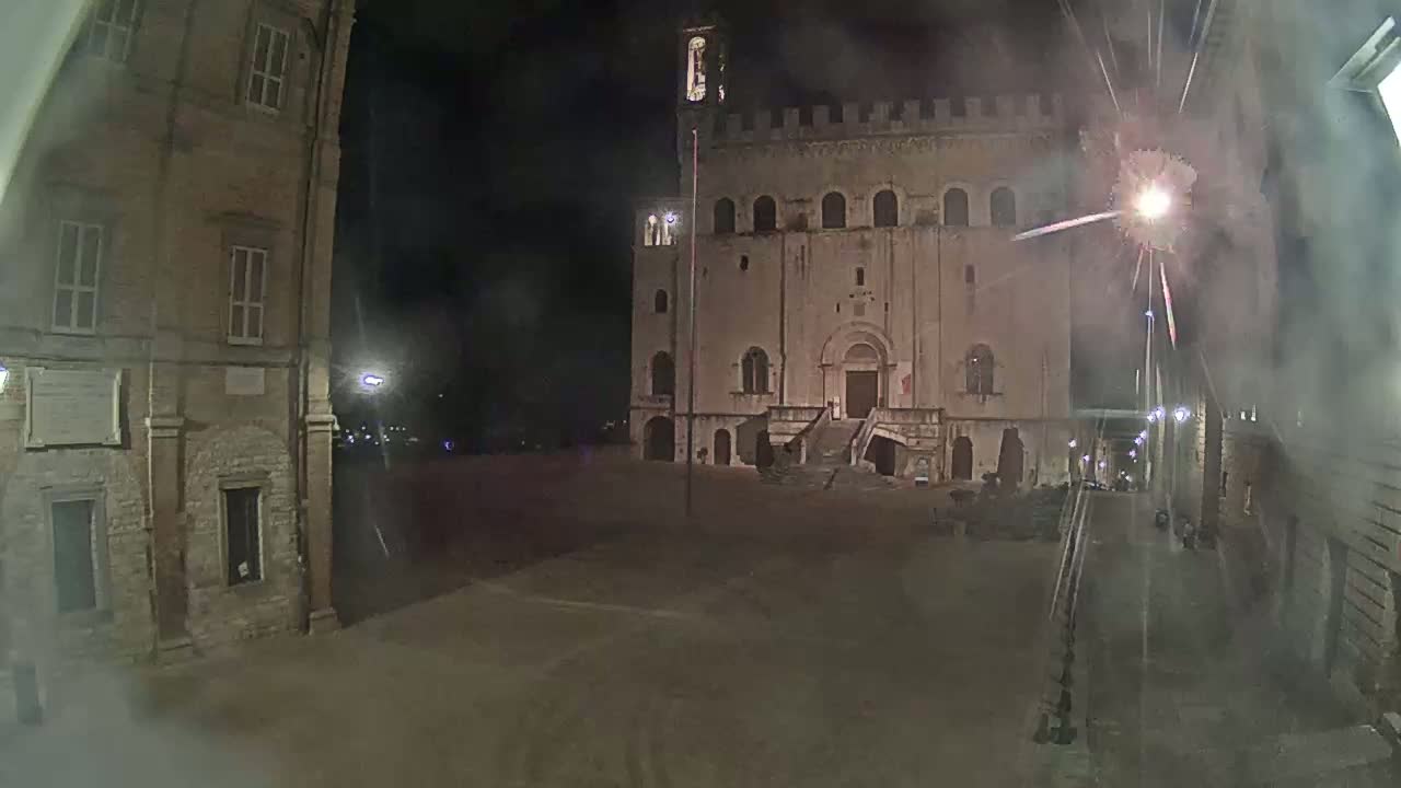 A large, stone building with a staircase stands in a dimly lit square at night, under a hazy sky with fireworks in the background.