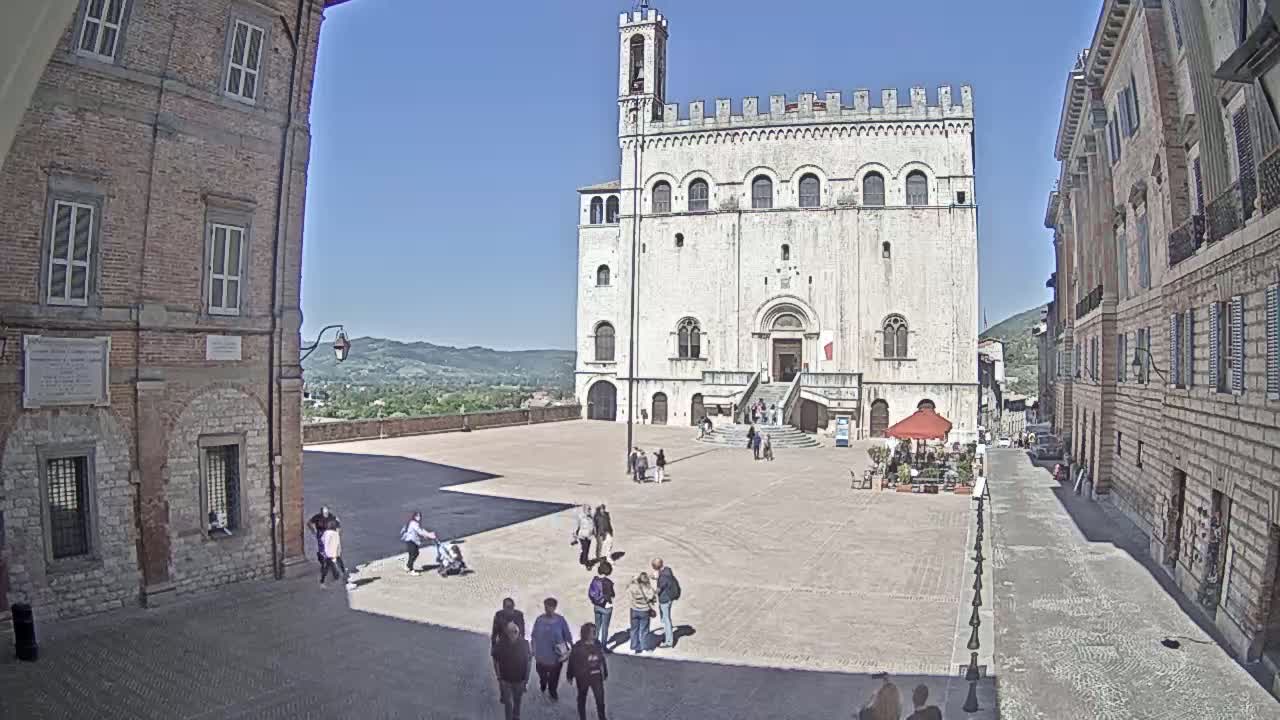 A large, light-colored stone building with a tower stands in a town square on a sunny day, with several people walking around and buildings lining a street leading away from the square.