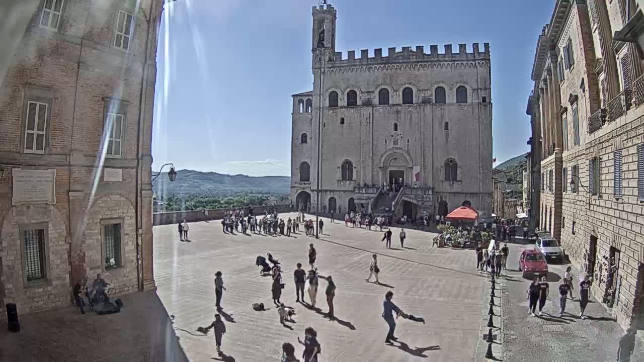 A large, light-colored stone building stands in a sunny town square, where people stroll and gather under a clear blue sky.