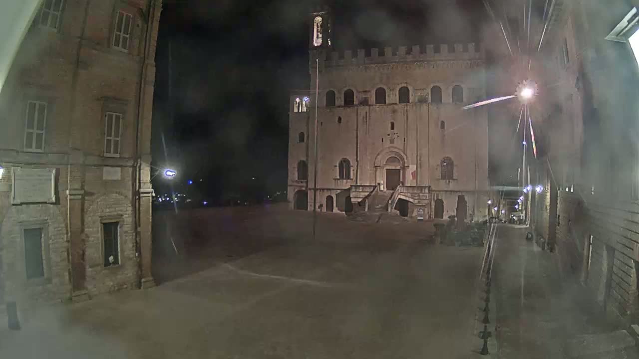 A large, stone building with a staircase stands in a dimly lit square at night, under hazy conditions.