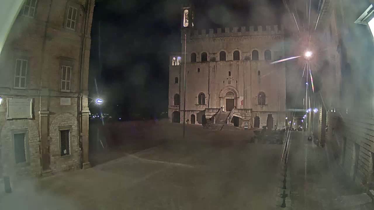 A large stone building with a staircase stands in a dimly lit square at night, with hazy weather conditions.