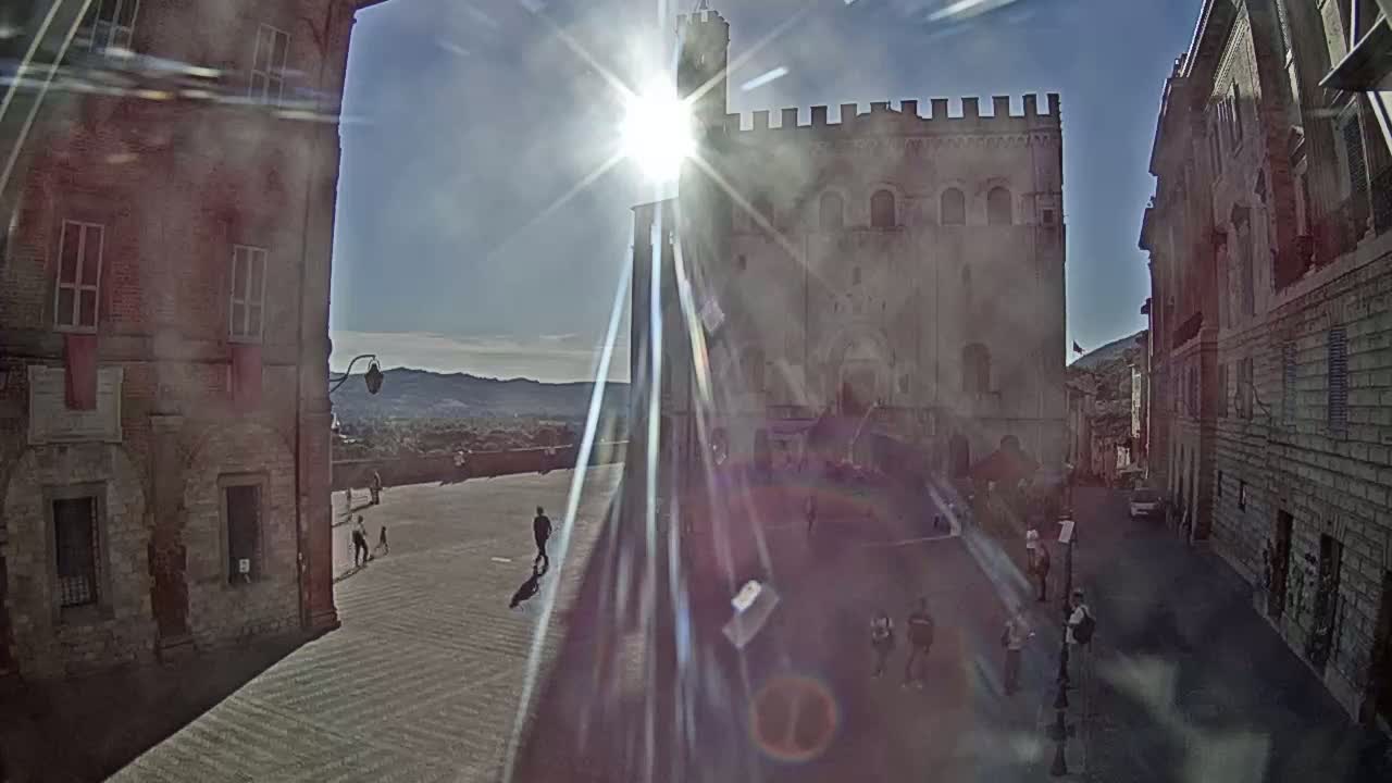 A sunny day reveals a town square surrounded by ancient stone buildings, with a few people strolling about.
