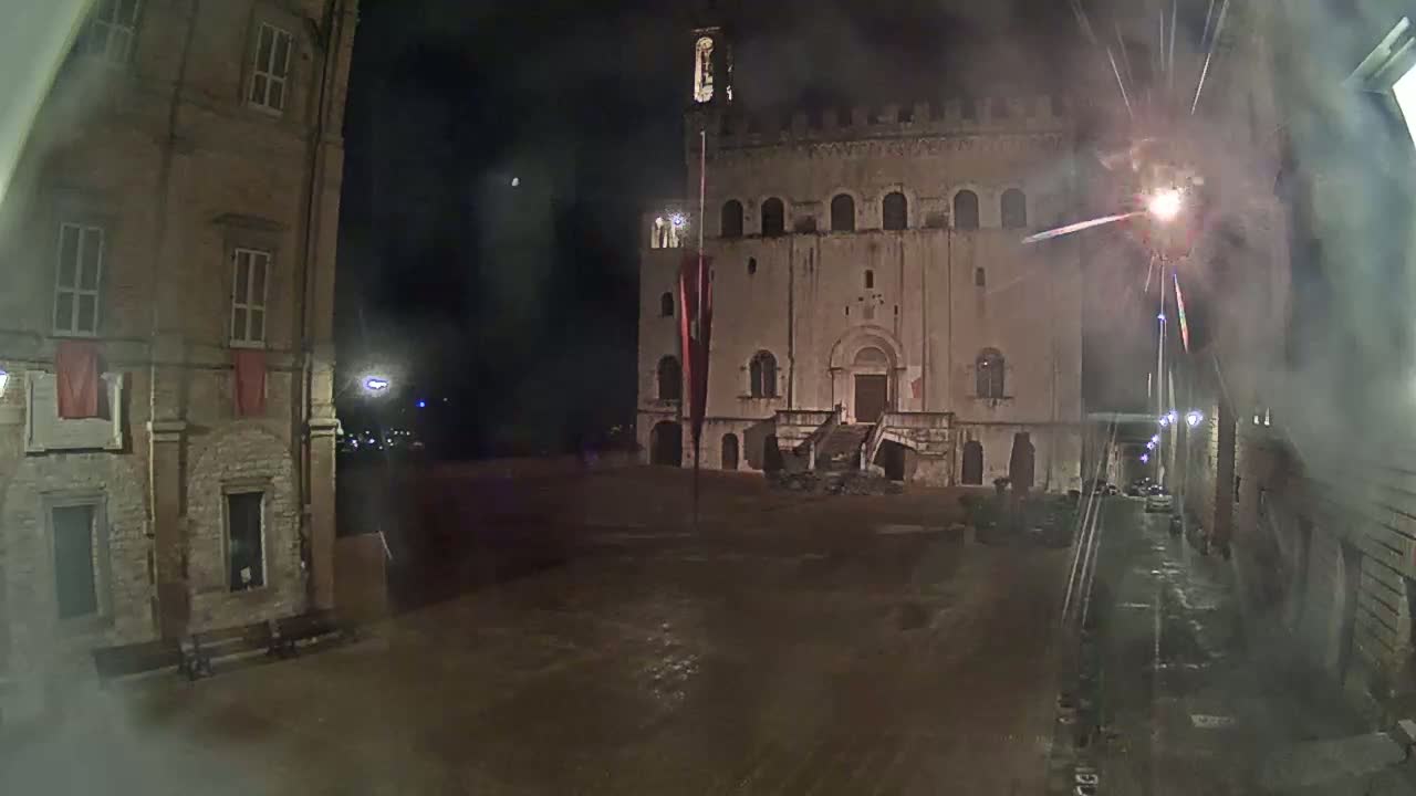 A nighttime view of a large stone building with fireworks exploding in the background, in a square between two buildings.