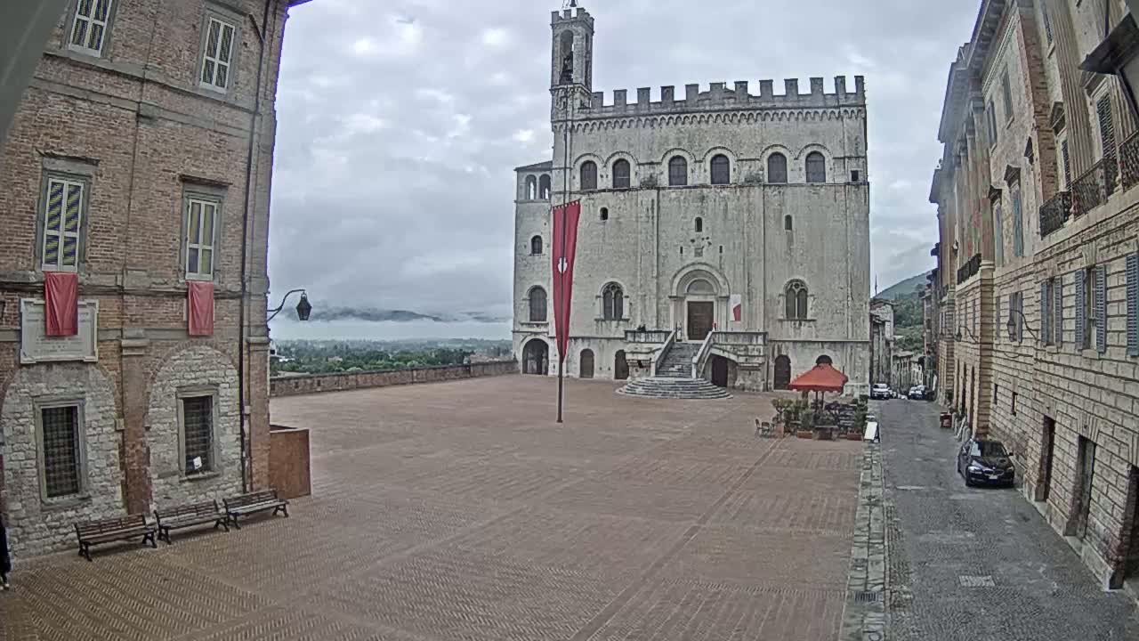 A large, stone edifice stands in a brick plaza between two brick buildings on a cloudy day, with a distant, misty landscape visible in the background.