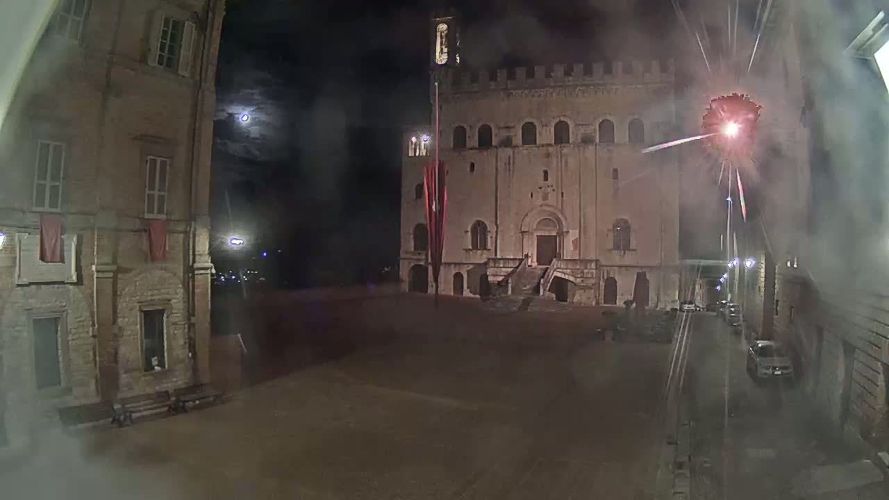 A large stone building with a staircase is visible at night under a partly cloudy sky, with fireworks exploding in the background.