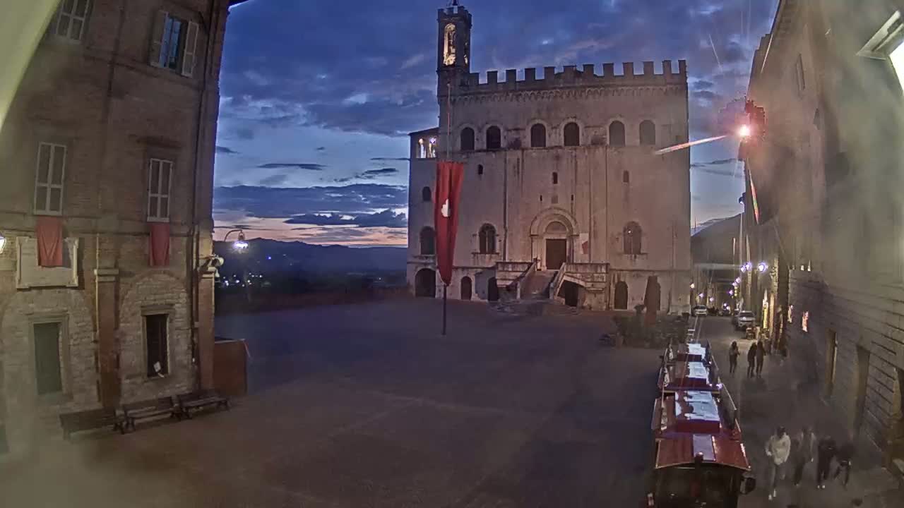 A large stone building with a tower stands in a town square at twilight, under a partly cloudy sky.