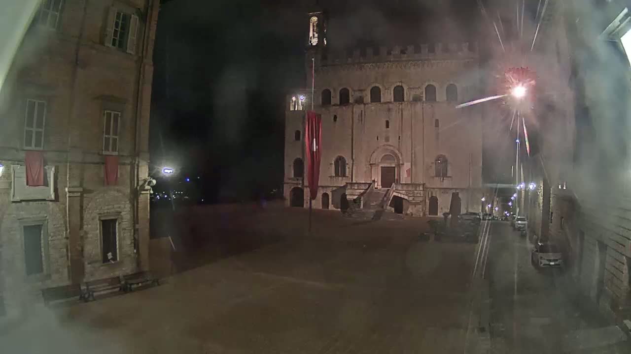 A nighttime view of a town square with a large stone building at the far end, illuminated by fireworks in a hazy atmosphere.