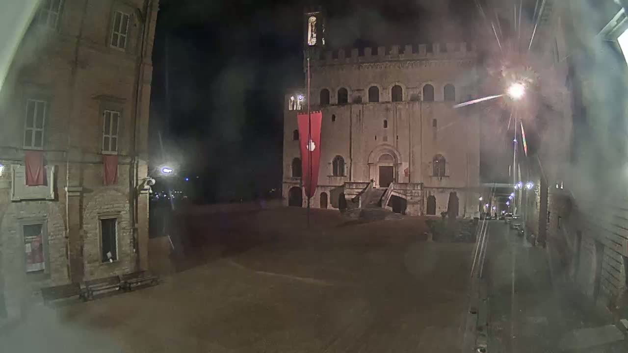 A nighttime view of a town square, featuring a large stone building with a red banner, flanked by older buildings under hazy or smoky conditions.