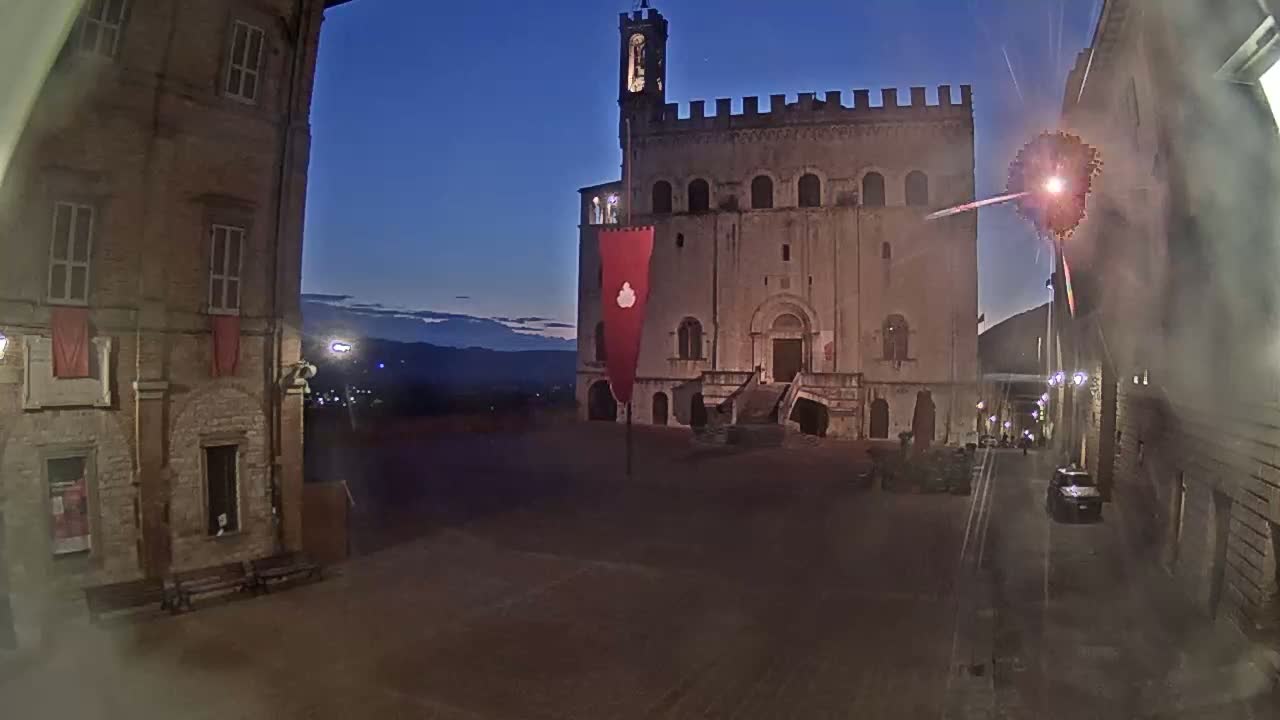 A large, stone building with a tower stands in a town square at twilight, under a clear, dark blue sky, with fireworks visible in the distance.