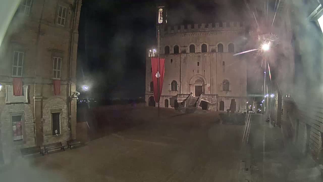 A nighttime view of a town square with fireworks, featuring a large stone building with a red banner and flanking buildings.