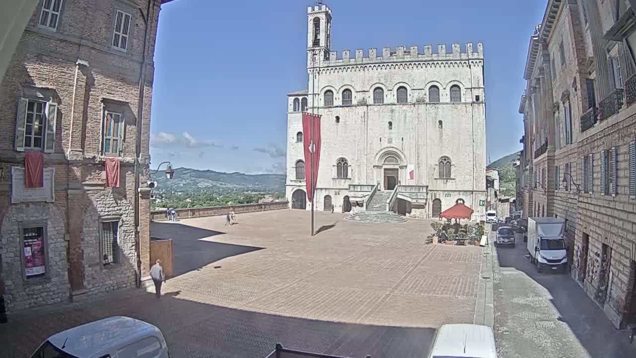 A large, light-colored stone building with a red flag stands in a town square on a sunny day, with other buildings lining the square and a view of rolling hills in the background.