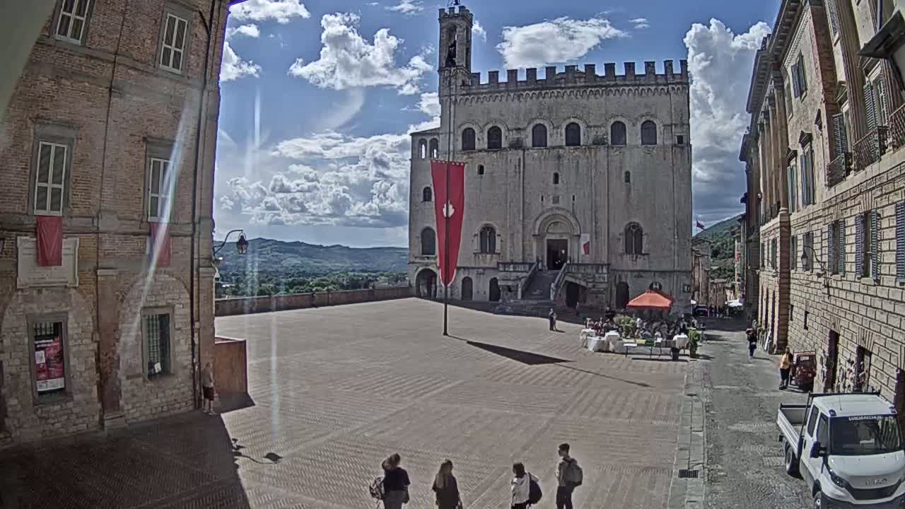 A large, light-colored stone building stands in a town square under a partly sunny sky, with several people walking in the square and others sitting at outdoor tables.