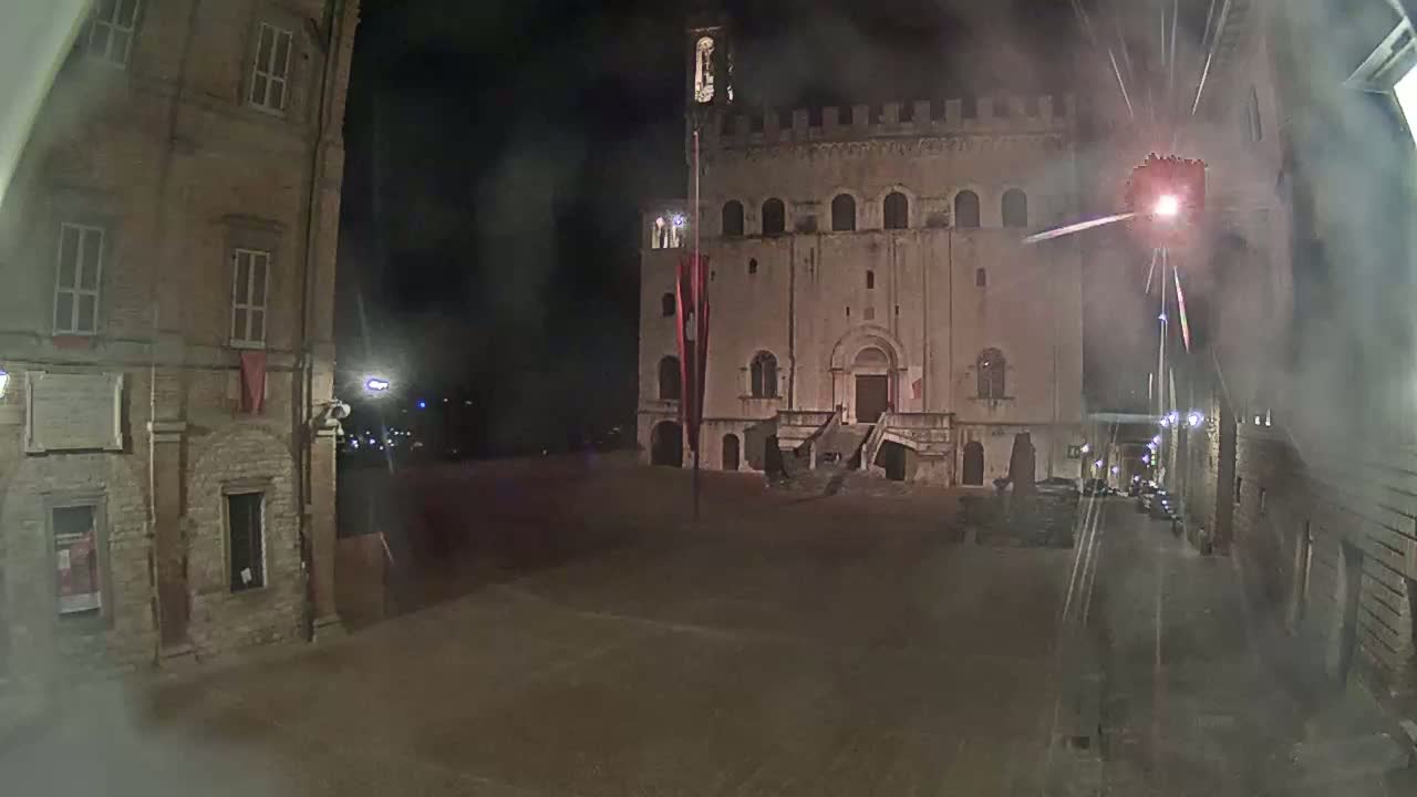 A large, stone building with a staircase stands in a dimly lit square at night, with a hazy atmosphere and faint lights in the distance.