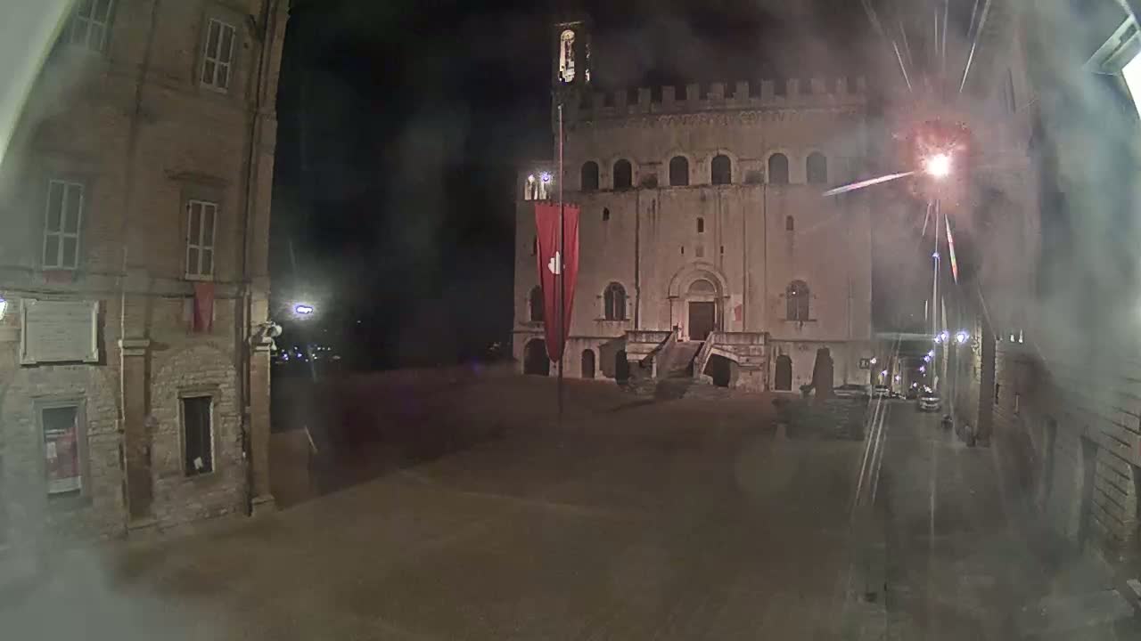 A large stone building with a flagpole and red banner stands in a dimly lit square at night, with fireworks exploding in the sky.