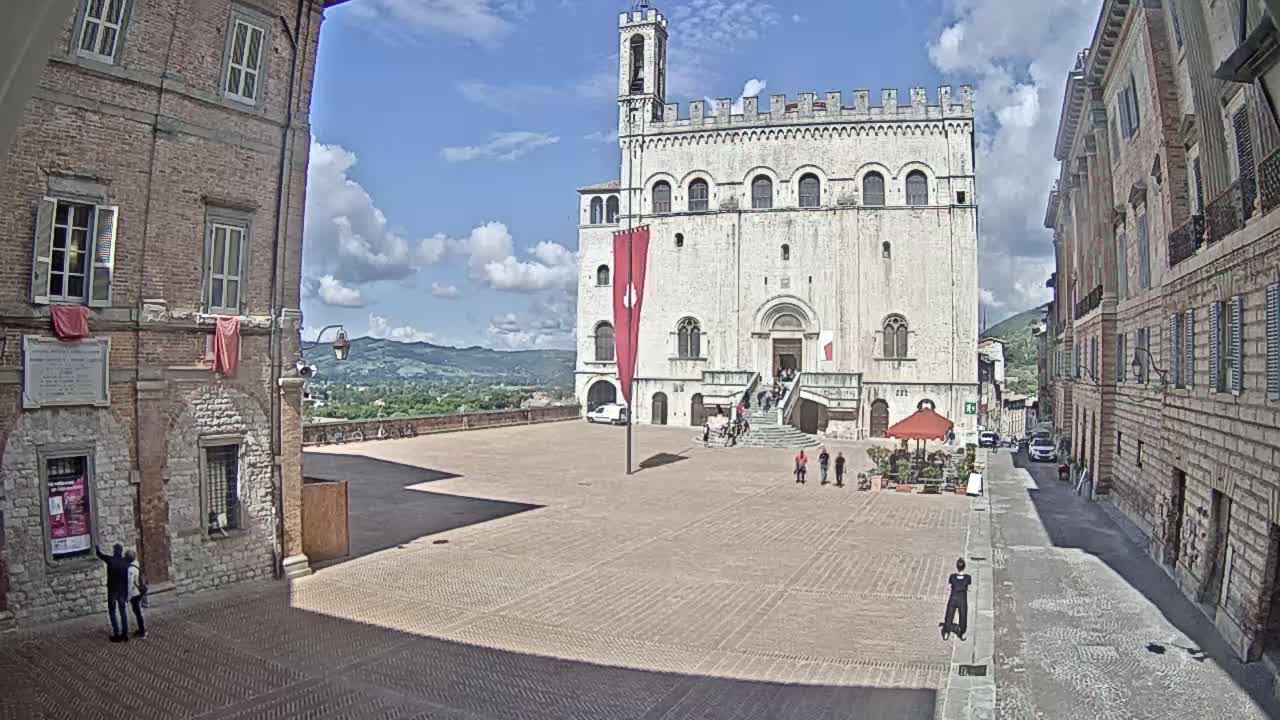 A large, light-colored stone building with a tall tower stands in a town square on a partly sunny day, flanked by brick buildings and overlooking distant hills.