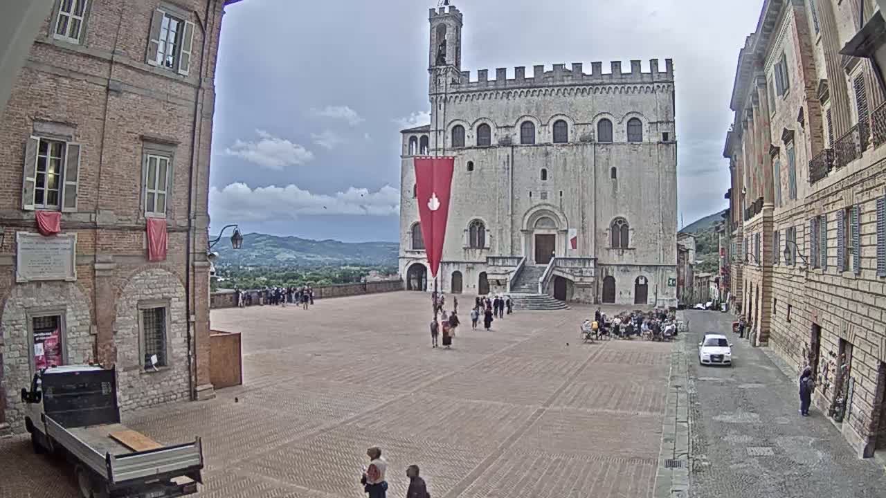 A large, light-colored stone building with a red banner stands in a town square, under a mostly cloudy sky, with people gathered in the square and along the surrounding buildings.