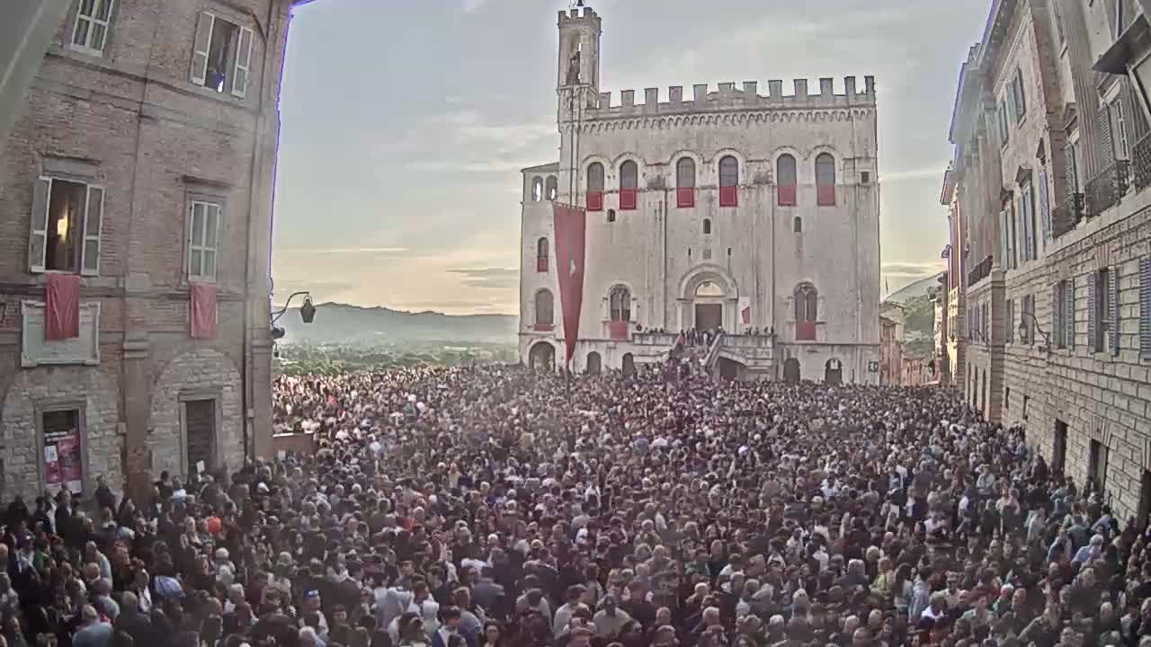 A large crowd fills a town square in front of a tall, light-colored building under a partly cloudy sky.