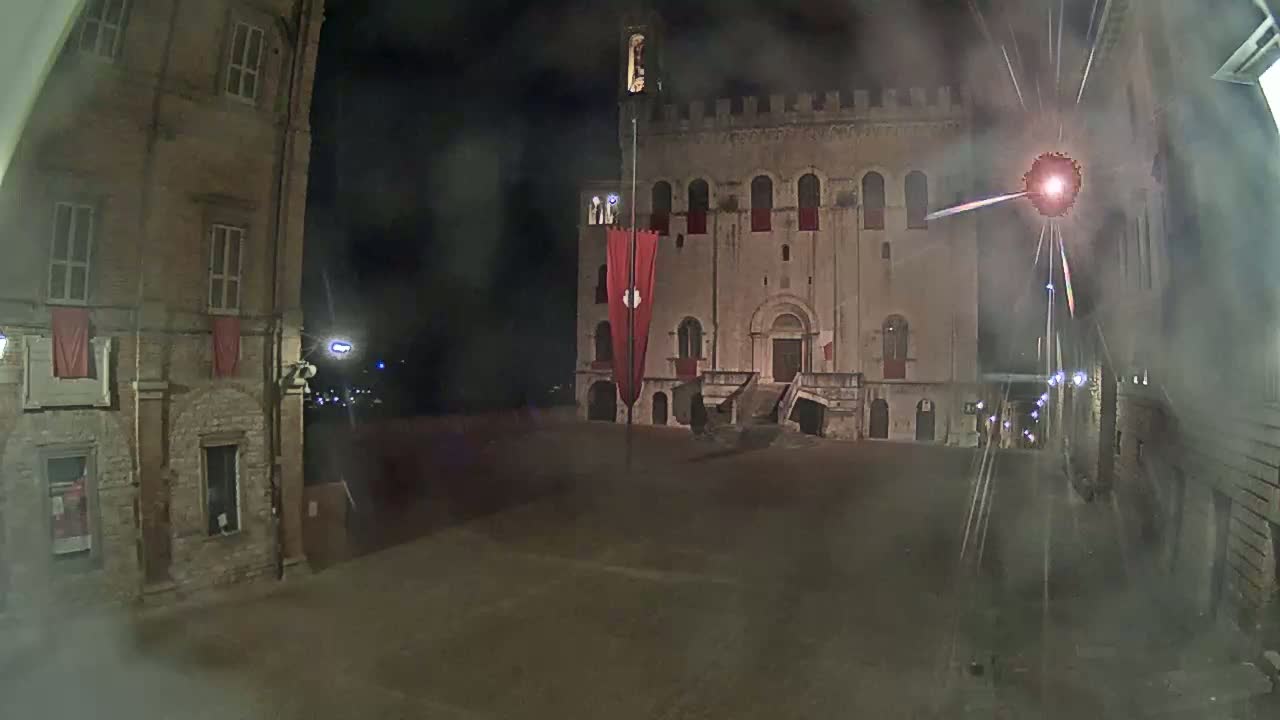 A large, stone building with a red banner stands in a mostly empty square at night, with a hazy or foggy atmosphere.