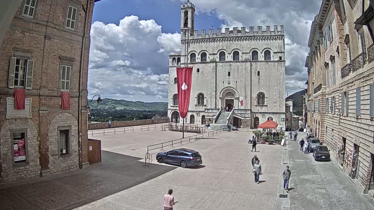 A large, light-colored stone building stands in a town square under a partly cloudy sky, with people walking around and cars parked nearby.