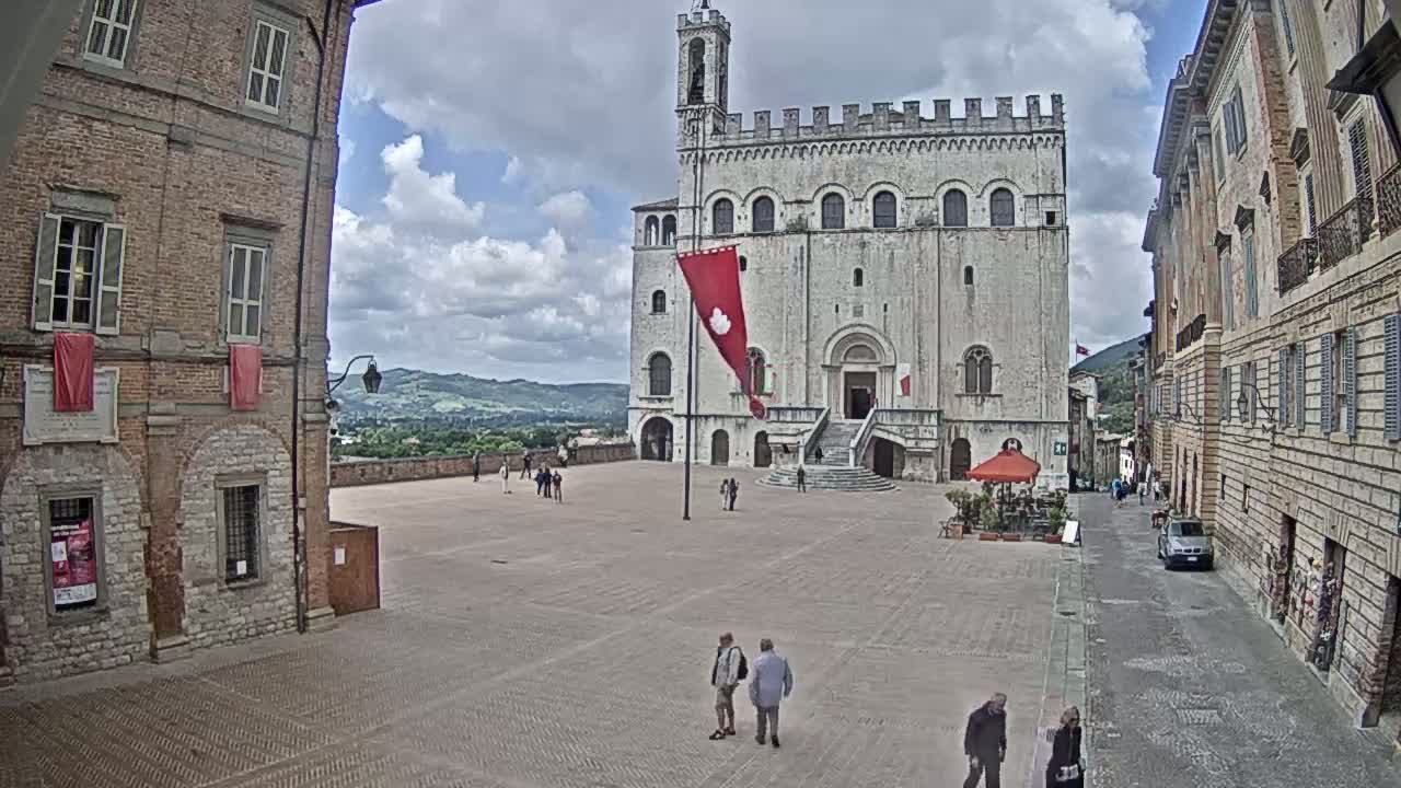 A large, pale stone building with a red flag stands in a town square on a partly cloudy day, with several people walking in the square and streets leading to it.