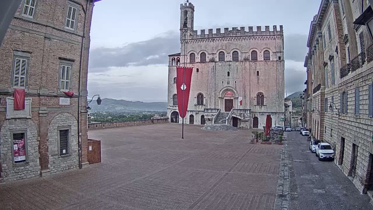 A large stone plaza, under an overcast sky, features a prominent stone building with a red banner, flanked by older stone buildings, and several cars parked on a narrow street.