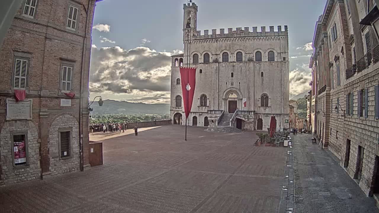 A large, light-stone public building with a red flag stands in a town square, surrounded by brick buildings under a partly cloudy sky.
