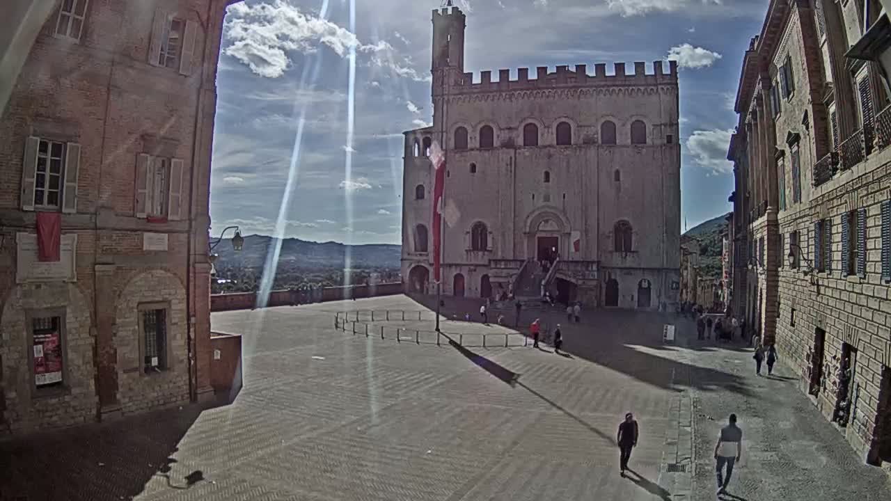A large, light-colored stone building with crenellations stands in a sunny town square, surrounded by other buildings and a few pedestrians.