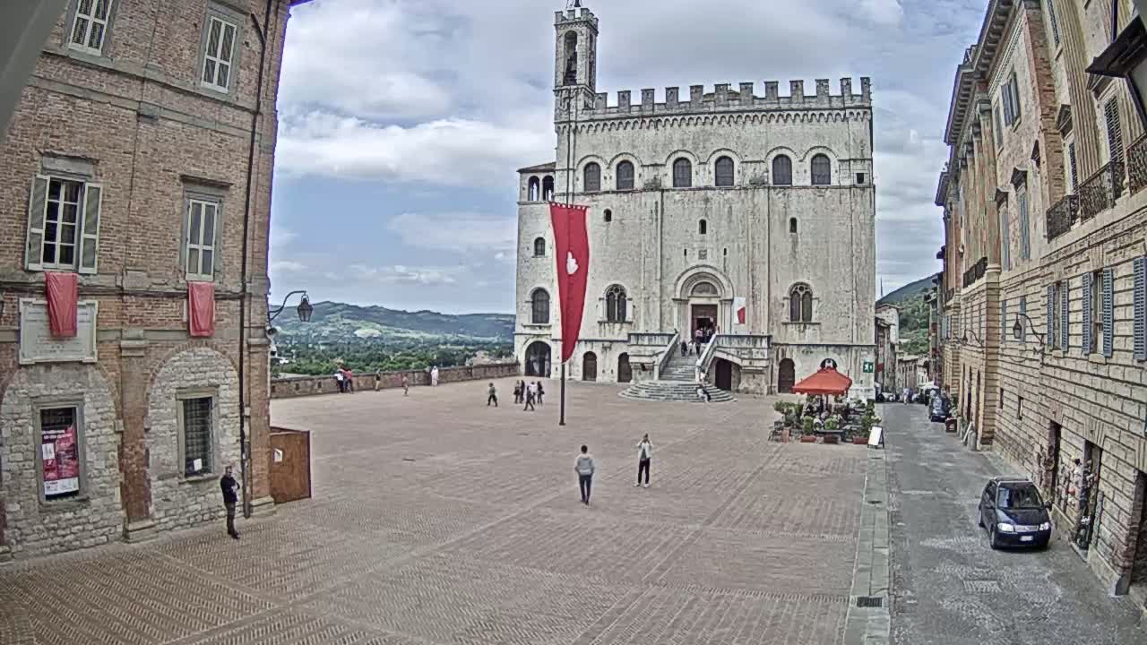 A large, light-colored stone building with a staircase and a red flag stands in a town square under a partly cloudy sky, flanked by brick buildings and a few people.