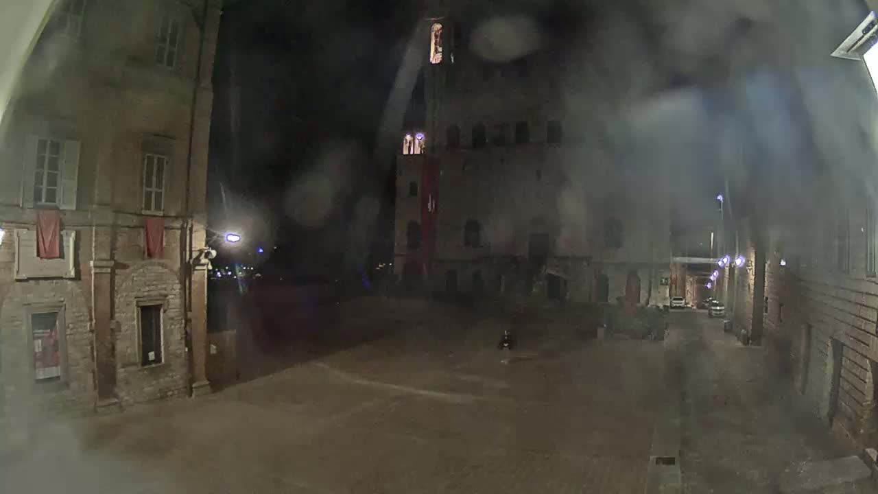 A rain-streaked nighttime view of a dimly lit stone plaza with buildings and a tall structure in the background.