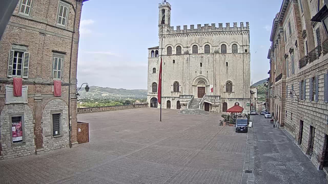A large, light-colored stone building with a staircase stands in a town square, flanked by brick buildings, under a mostly clear sky.