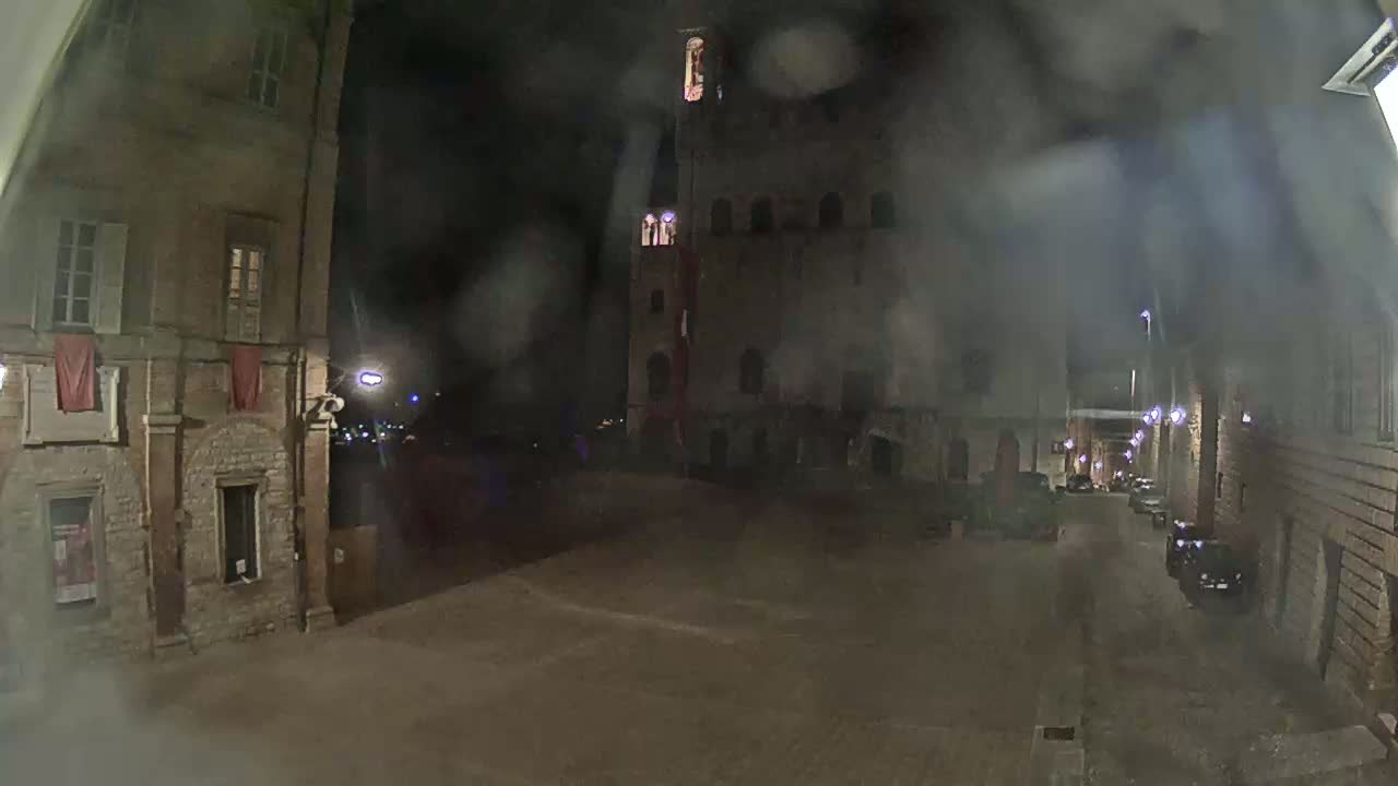 A nighttime view of a dimly lit, cobblestone town square, flanked by old stone buildings under a hazy, possibly rainy sky.