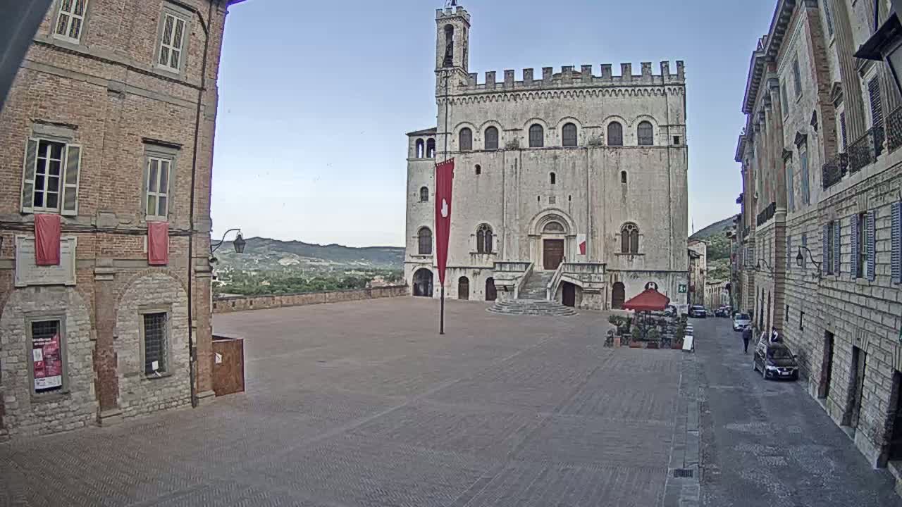 A large, light-colored stone building with a red banner stands in a town square on a clear day, flanked by brick buildings and with a few cars parked on a nearby street.