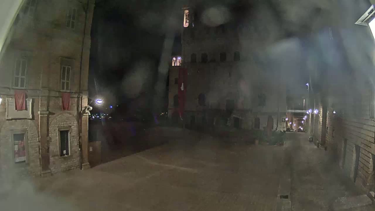 A rain-streaked nighttime view shows a square between stone buildings, one of which is a tall structure with multiple windows.