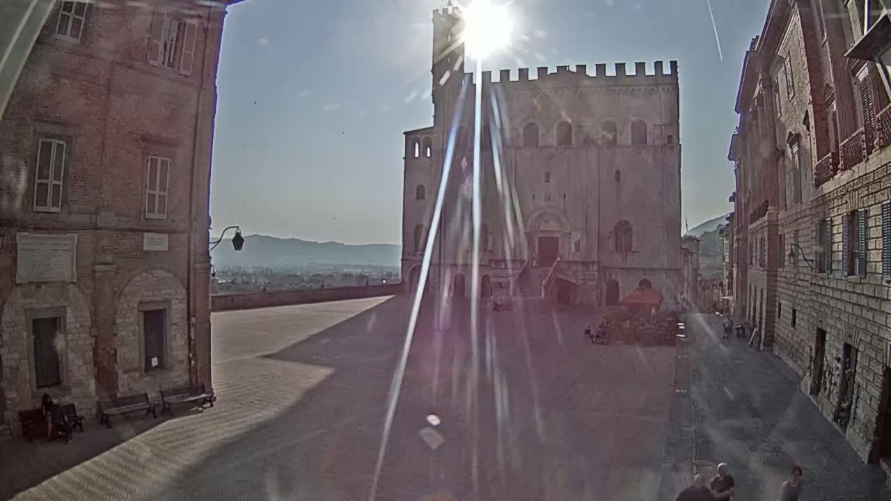 A sunny day reveals a large plaza with ancient stone buildings, one prominently featuring a tower, overlooking a distant valley.