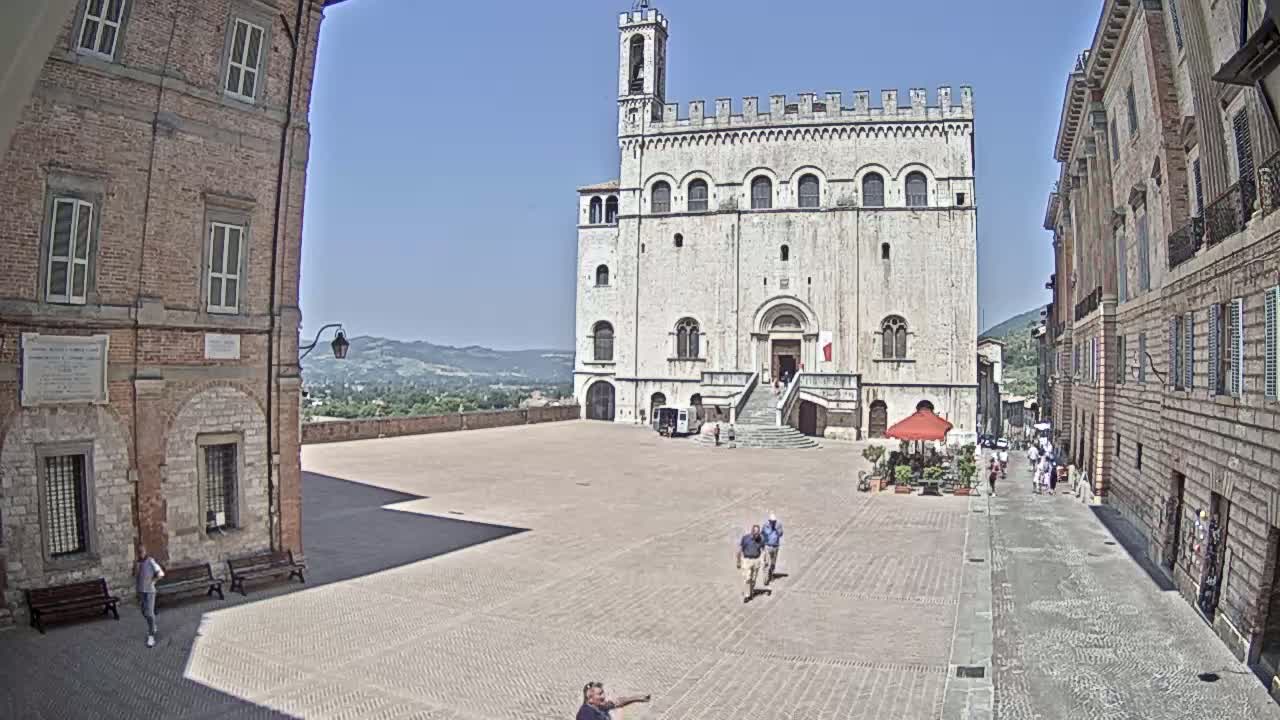 A large, light-colored stone building stands in a town square on a sunny day, with brick buildings lining the square and hills visible in the distance.