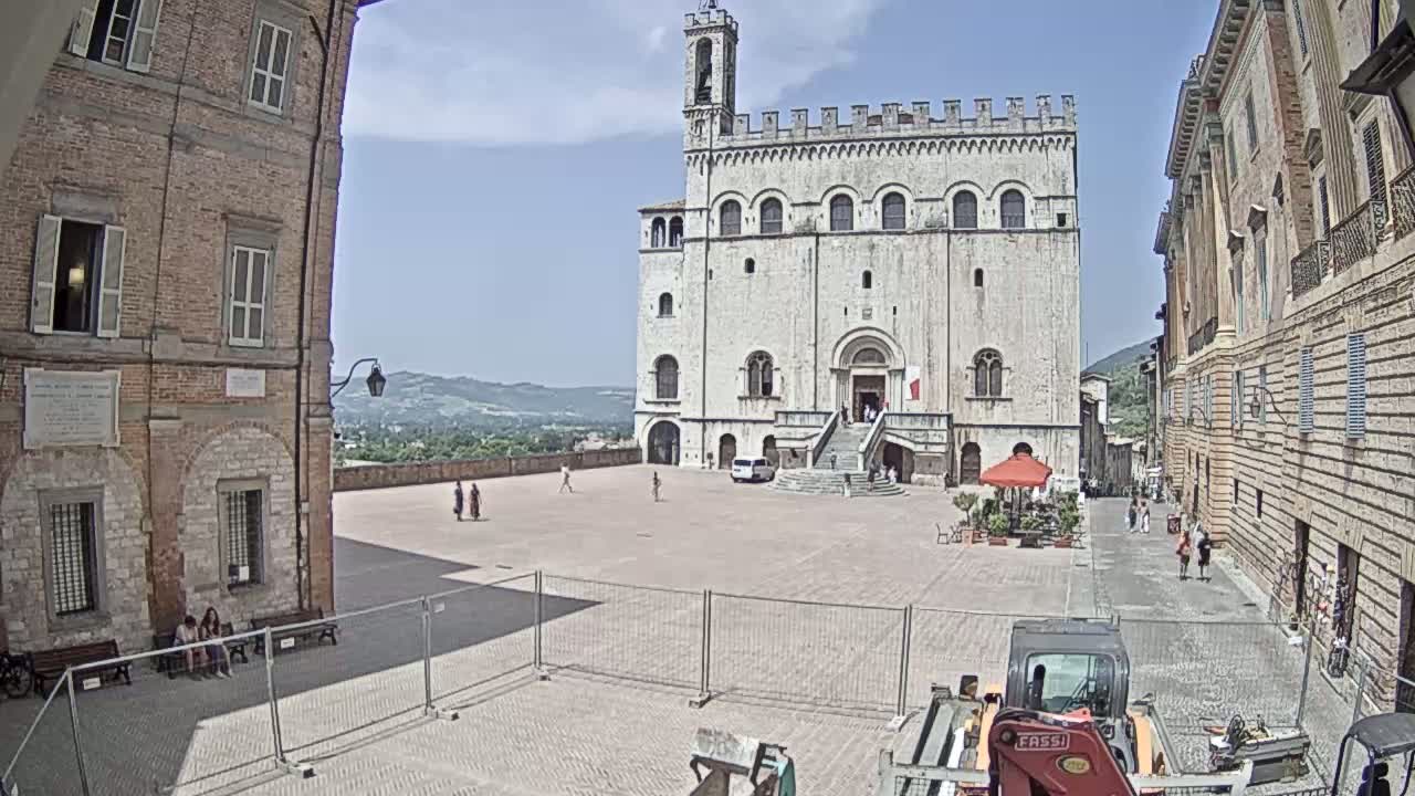 A large, light-colored stone building with a staircase stands in a mostly empty town square on a sunny day, flanked by brick buildings and a small amount of construction equipment.