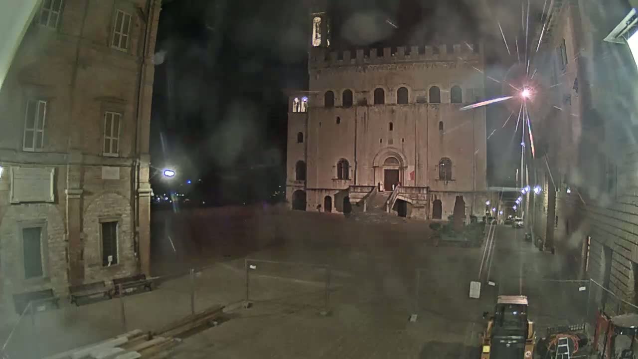 A large stone building with a staircase is visible in a nighttime square under a slightly hazy sky with fireworks exploding in the distance, construction equipment is present in the foreground.
