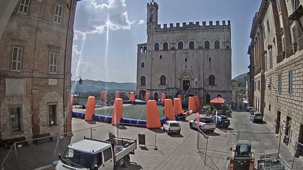 A large, light-colored stone building stands in a town square on a sunny day, with an inflatable structure in the foreground and several cars parked nearby.