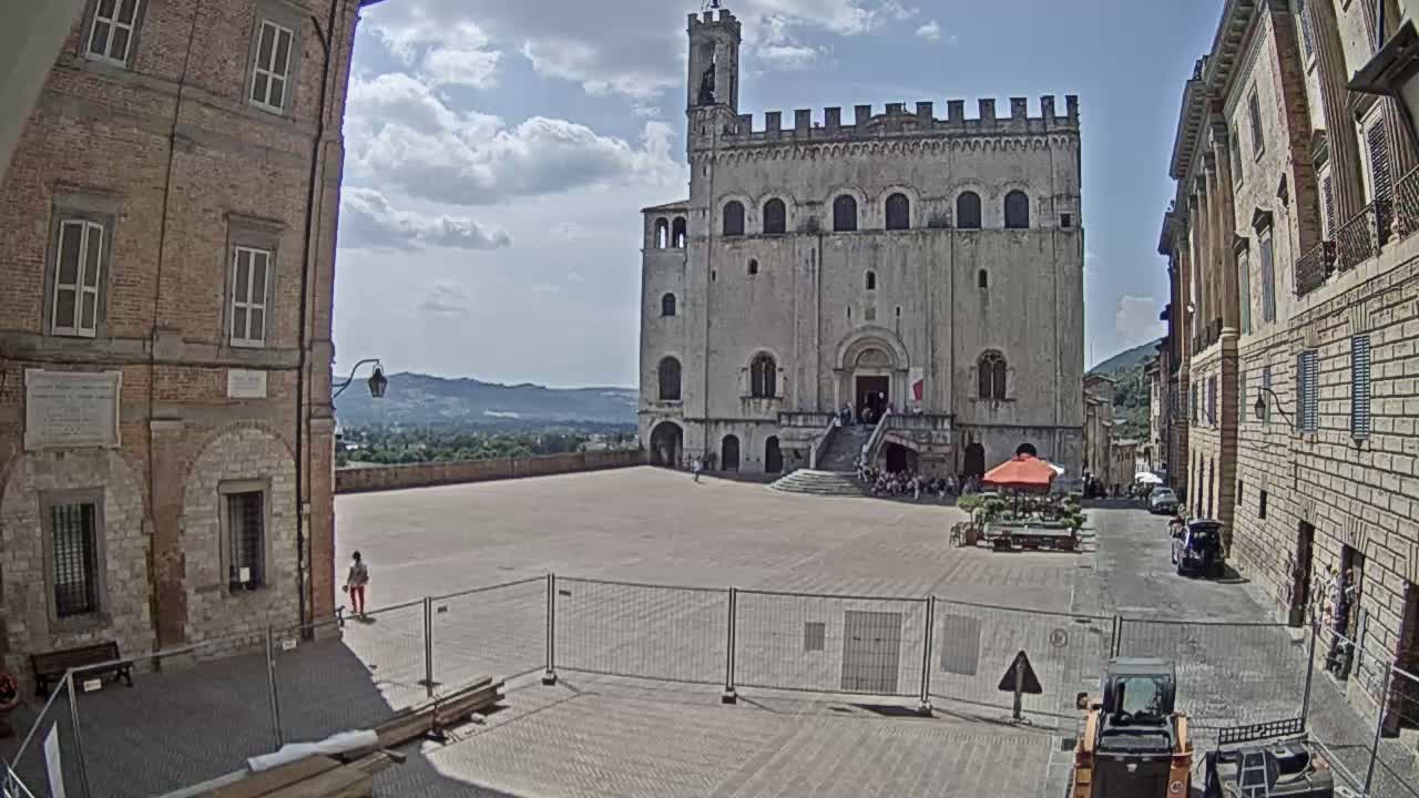 A large, light-colored stone building stands in a town square on a partly cloudy day, surrounded by other buildings, with some people visible and construction equipment present.