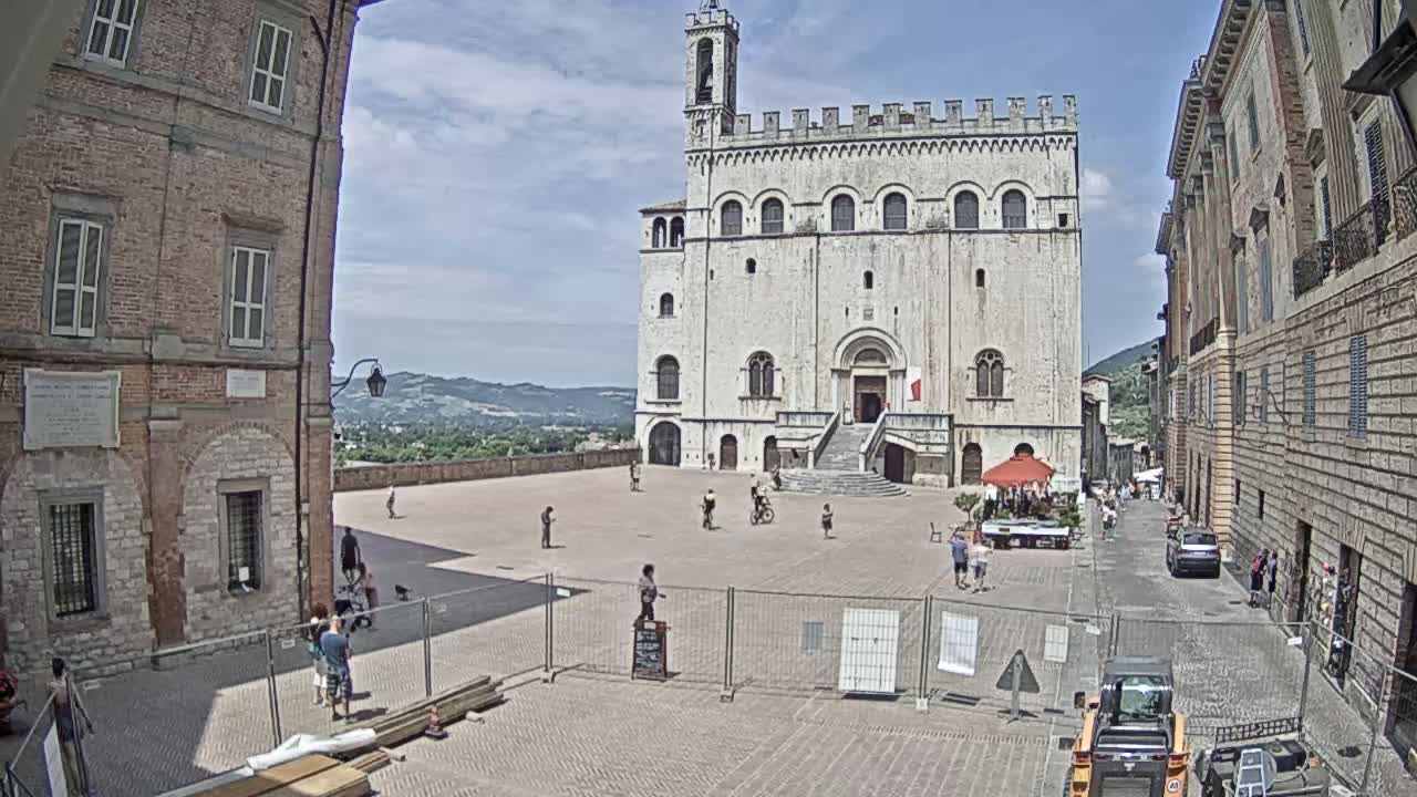 A large, light-colored stone building with a staircase stands in a town square on a sunny day, surrounded by brick buildings and a few people.