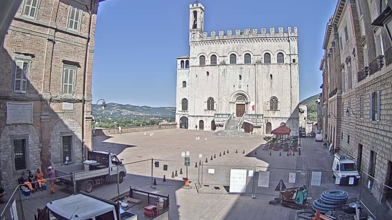 A large, light-colored stone building with a staircase stands in a town square on a sunny day, surrounded by other buildings and some construction equipment.