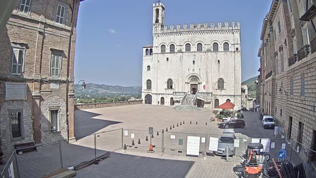 A large, light-colored stone building with a tower stands in a town square on a sunny day, with brick buildings flanking it and construction barriers in the foreground.