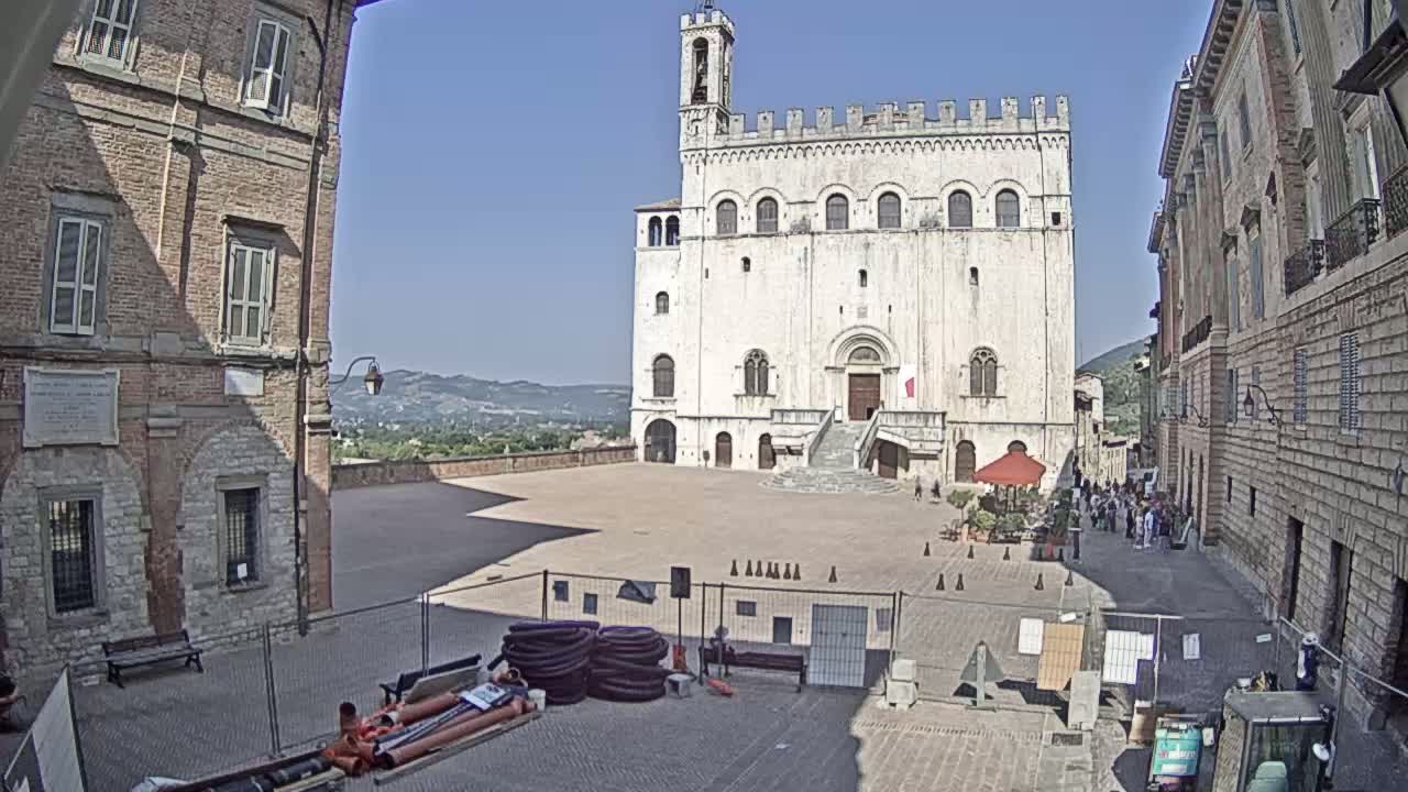 A large, light-colored stone building stands in a paved square on a sunny day, flanked by brick buildings and overlooking a distant valley.