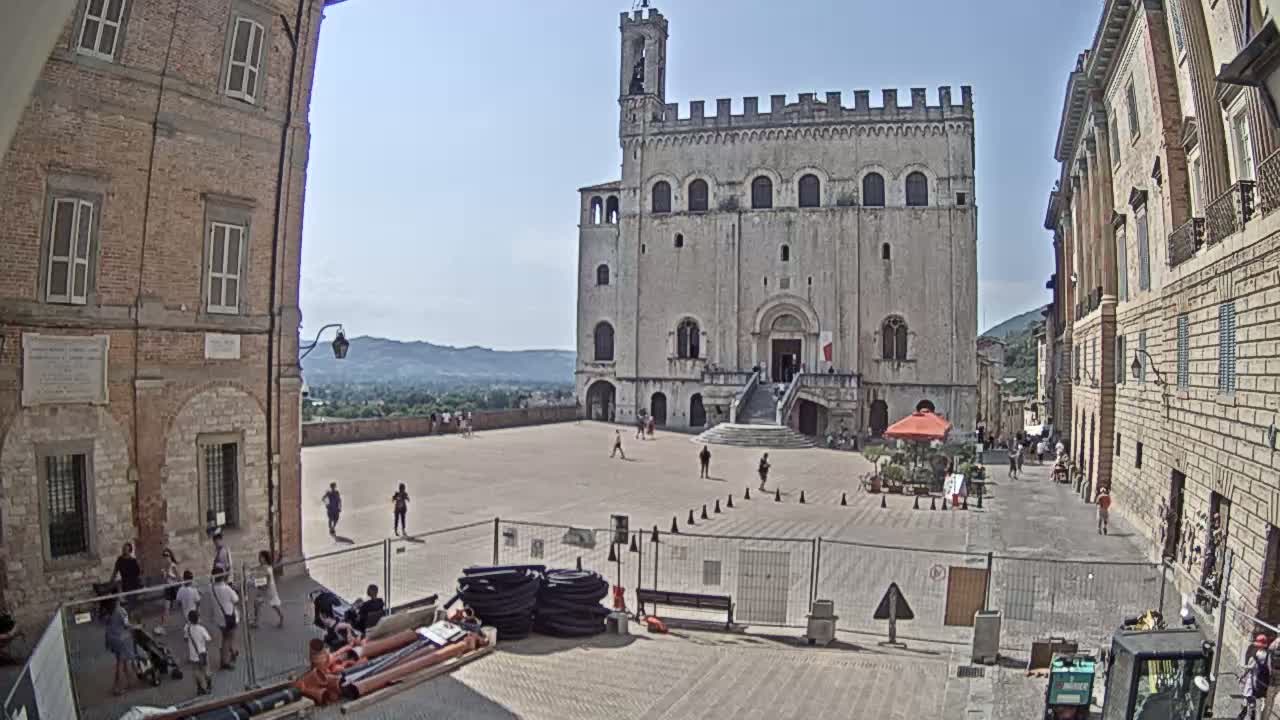 A large, light-colored stone building stands in a paved square on a sunny day, flanked by brick buildings and overlooking a distant valley.
