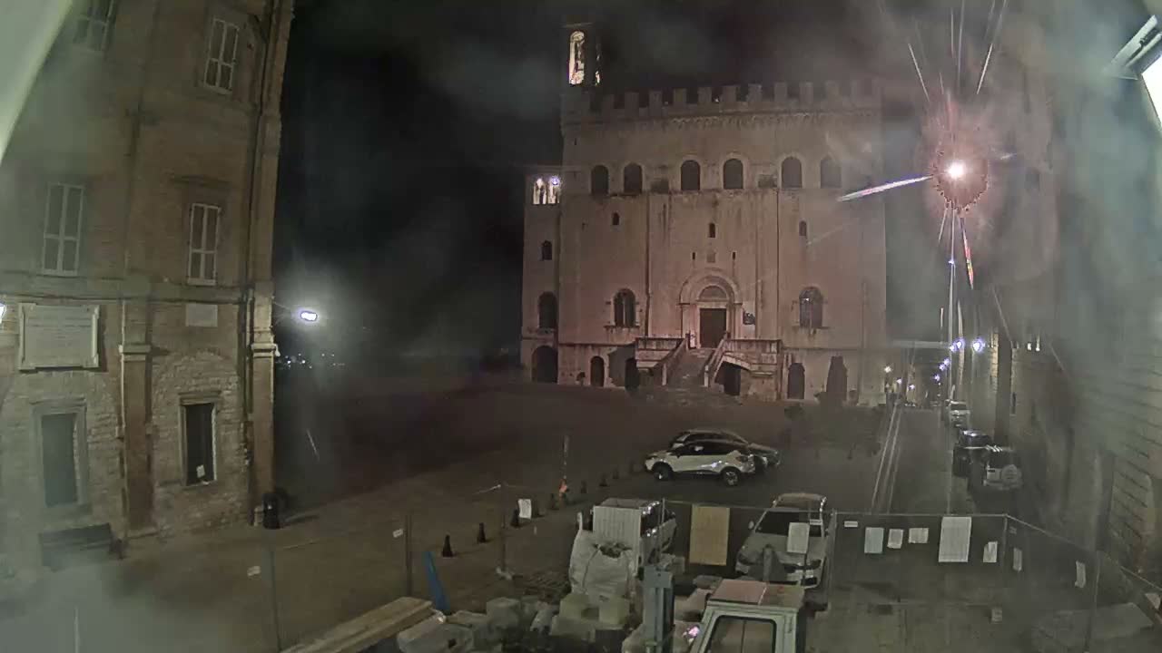 A historic town square is illuminated at night, featuring a large stone building with a bell tower and battlements, another brick building, parked cars, and construction materials in the foreground, all under a clear night sky.