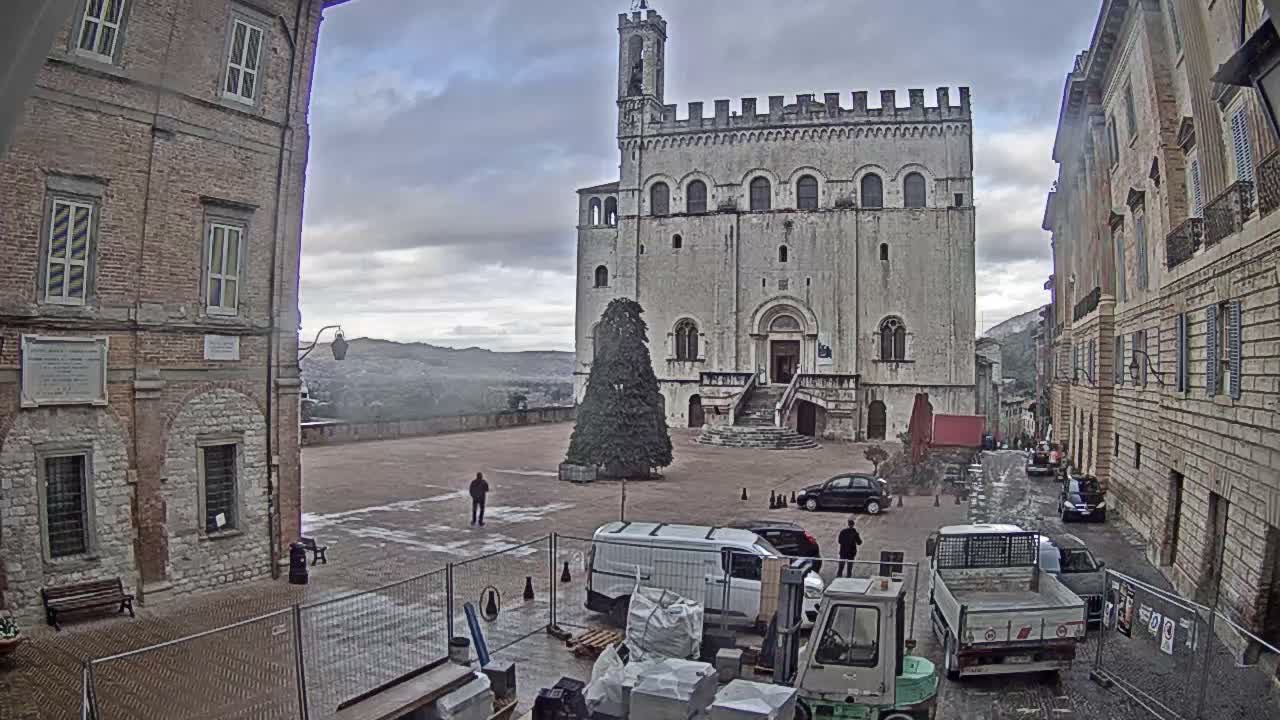 Heavily overcast skies loom over a historic European town square, which features a grand medieval-style stone building with a bell tower, a tall evergreen tree, and a busy construction area with vehicles and materials in the foreground, flanked by older brick buildings and distant hazy hills.