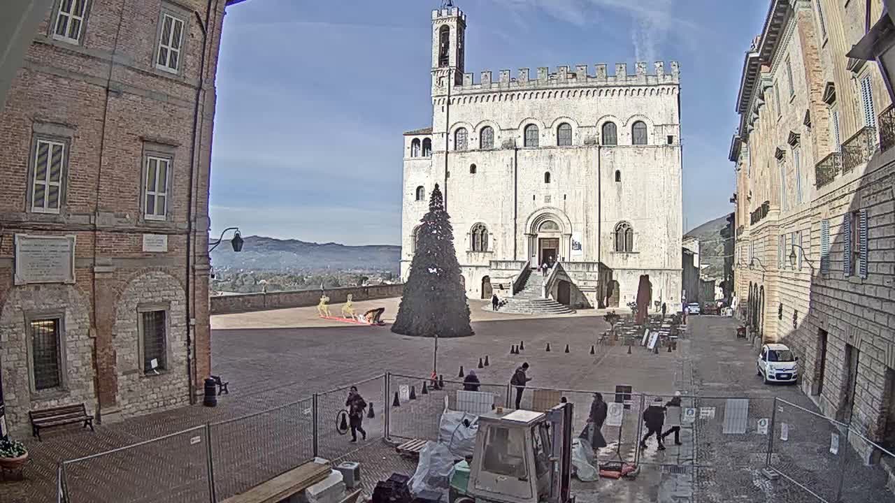 On a clear, sunny day, a historic town square is depicted with a grand stone building featuring a bell tower, a large Christmas tree, decorative reindeer, and various people moving around amidst foreground construction.