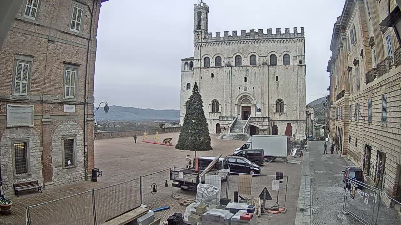 A grand historic stone building with a bell tower and battlements dominates a town square on an overcast day, featuring a large undecorated evergreen tree, various vehicles, scattered people, and foreground construction work, all set against distant hills.